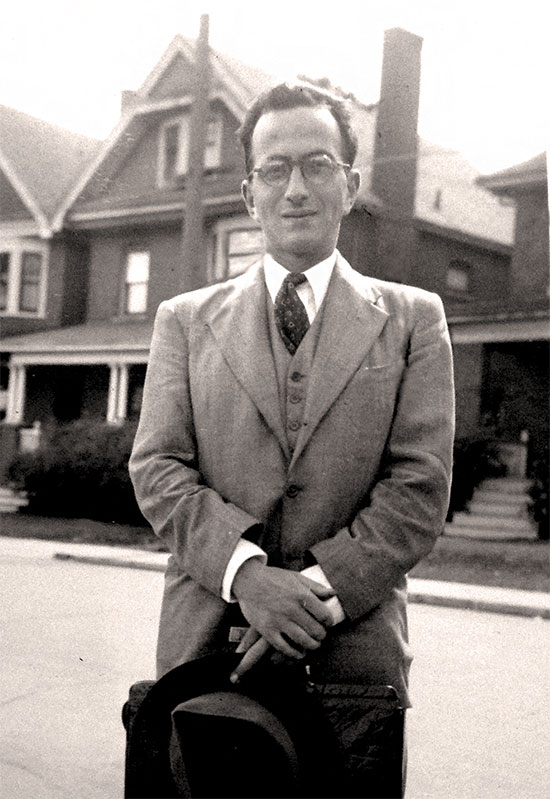 Black and white photo of Emil Fackenheim standing on a street in front of a row of houses.
