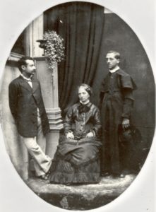 Black and white archival photo of a man in priest's robes standing next to a woman, seated, and a second man, standing. 
