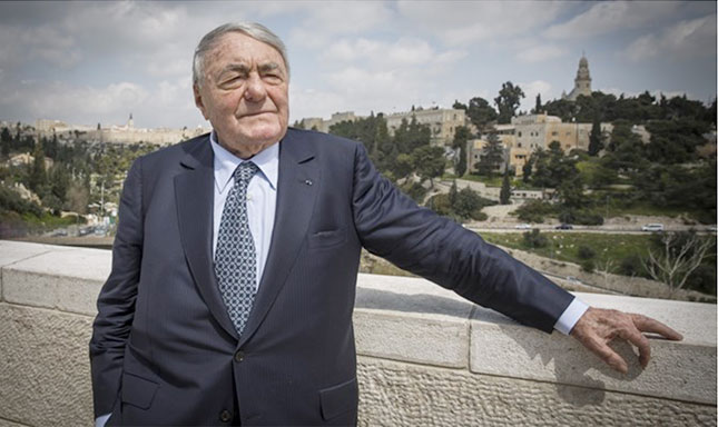 Photograph of a man in a suit leaning against a wall with Israel landscape in the background.