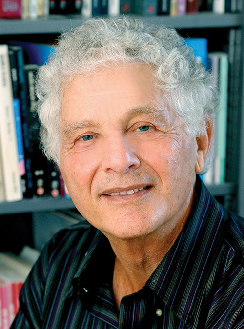 Photo of gray-haired smiling man in front of bookshelf.