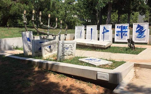 The aftermath of the 2017 attack on the memorial on Aristotle University's campus in Thessaloniki. Show slabs of marble off kilter and painted with Greek letters and a cross.