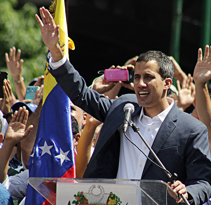 Juan Guaidó addresses a February 2019 protest in Caracas. The photo show a man in front of a microphone, raising his hand. A crowd of people behind him also have their hands raised. 