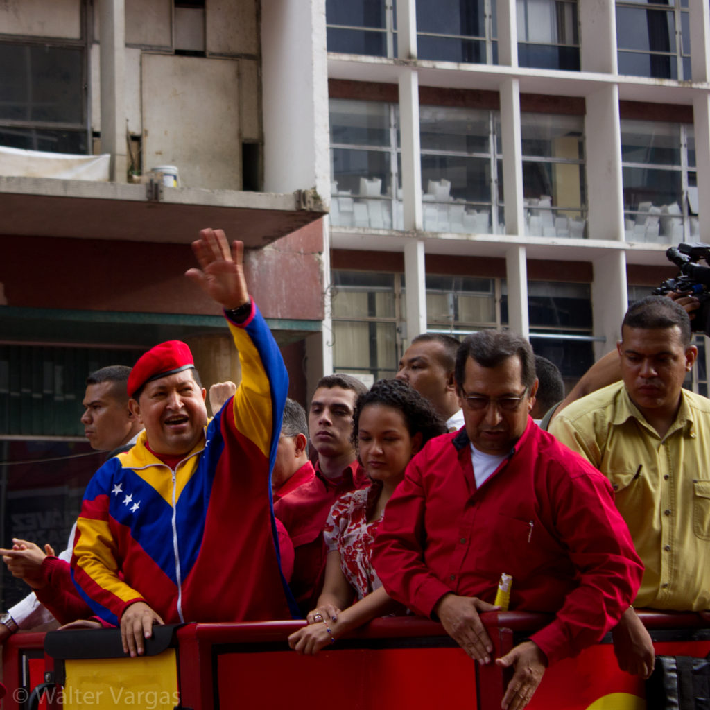 Hugo Chavez, in track suit that uses the colors and shapes of the flag of Venezuela and a military beret, waves to a crowd.